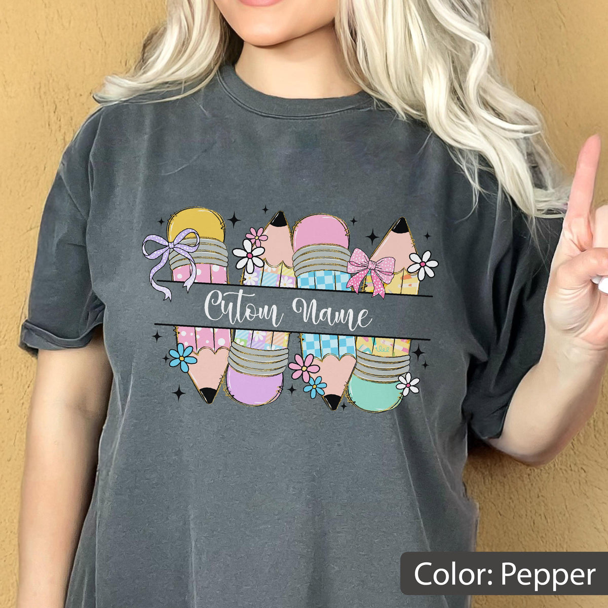 May include: A charcoal gray t-shirt featuring a graphic of four pencils in pastel shades of pink, blue, and yellow with checkered and floral patterns. The design includes small flowers, bows, and a central horizontal banner with the text Cutom Name.
