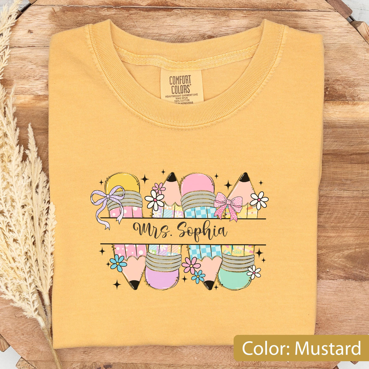May include: A mustard yellow t-shirt featuring a graphic print of multiple pencils decorated with bows, flowers, and checkered patterns. The text "Mrs. Sophia" is printed in a black script font across the center of the pencil design.