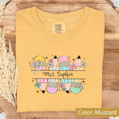 May include: A mustard yellow t-shirt featuring a graphic print of multiple pencils decorated with bows, flowers, and checkered patterns. The text "Mrs. Sophia" is printed in a black script font across the center of the pencil design.