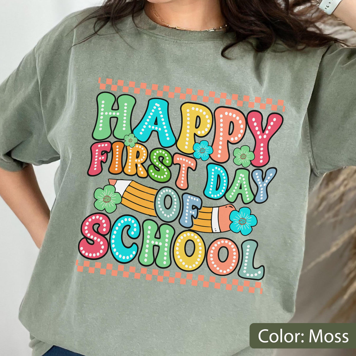May include: A moss green t-shirt featuring a colorful graphic with the text "HAPPY FIRST DAY OF SCHOOL" in a retro, bubbly font. The design includes a yellow pencil illustration, small flower accents, and a checkered border pattern.