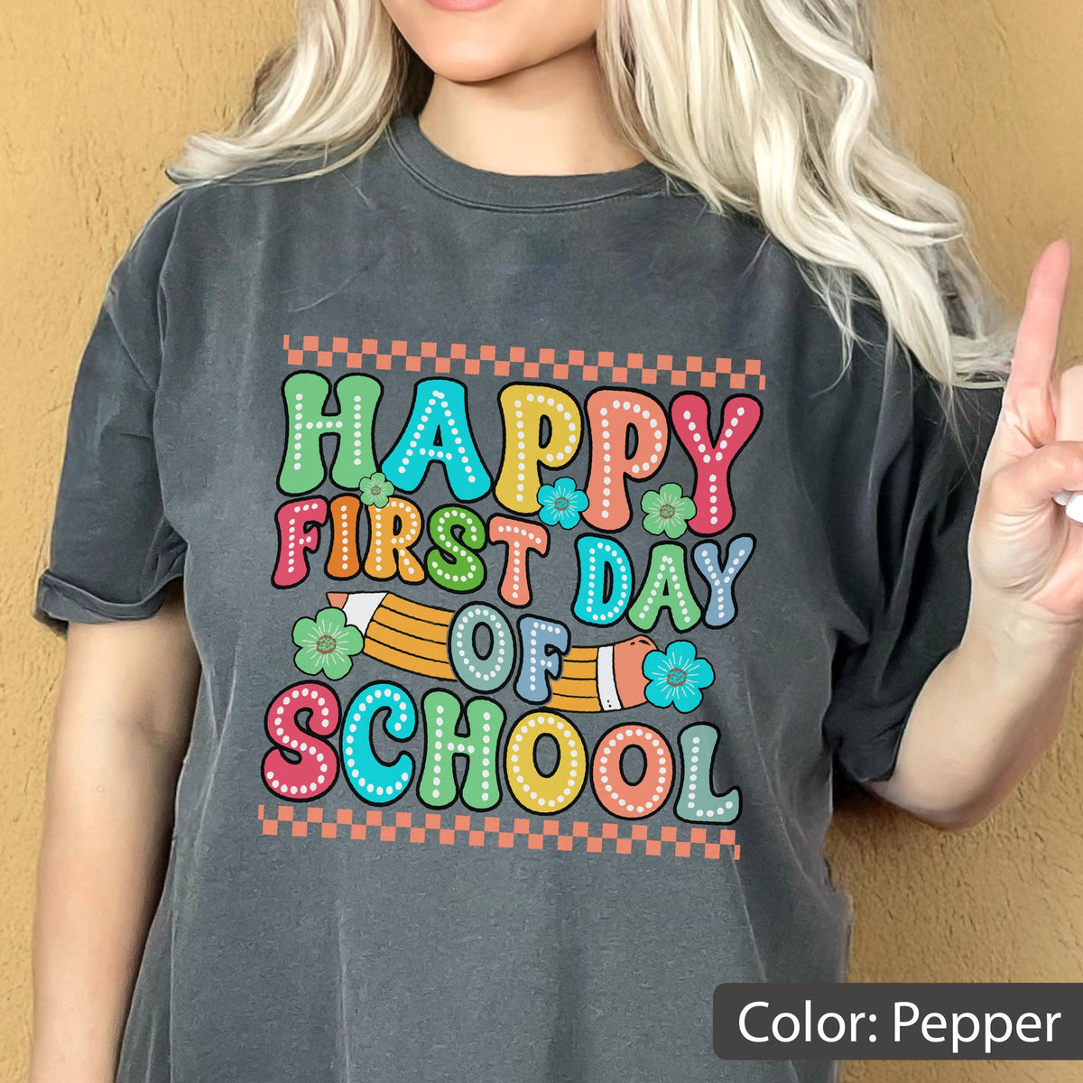 May include: A pepper-colored short-sleeve t-shirt featuring a graphic print with the text "HAPPY FIRST DAY OF SCHOOL" in colorful, bubbly letters. The design includes a yellow pencil illustration, small flowers, and orange checkered borders.