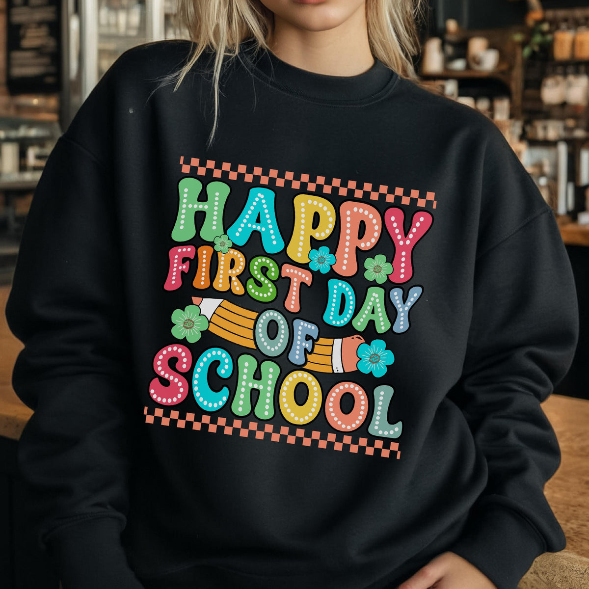 May include: A black crewneck sweatshirt featuring a colorful graphic print that reads Happy First Day Of School in a retro bubble font. The design includes a yellow pencil illustration, small green and blue flowers, and orange checkered borders.