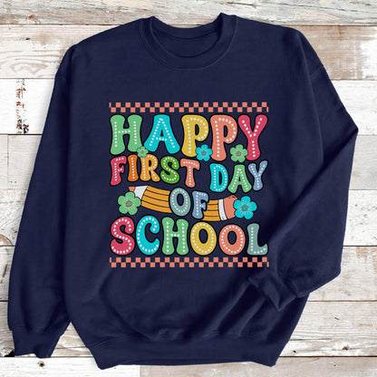 May include: A navy blue long-sleeve sweatshirt featuring a colorful graphic print that reads Happy First Day Of School in a dotted font. The design includes a yellow pencil illustration, small teal flowers, and orange checkered borders.