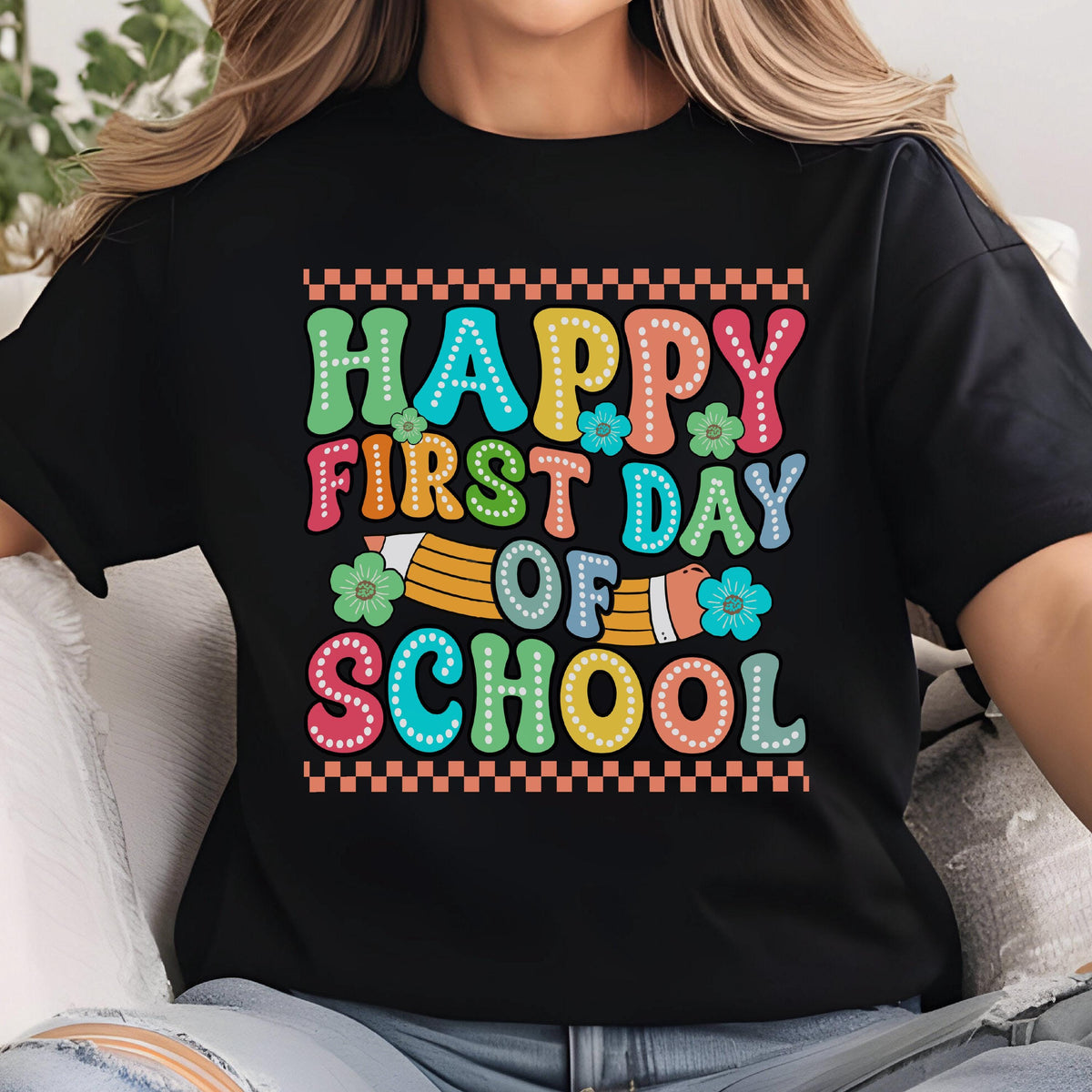 May include: A black t-shirt featuring the text HAPPY FIRST DAY OF SCHOOL in colorful, dotted, bubble-style letters. The design includes a yellow pencil graphic, small blue and green flowers, and a checkered border at the top and bottom.