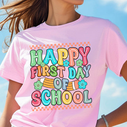 May include: A light pink short-sleeve t-shirt featuring a colorful graphic with the text "HAPPY FIRST DAY OF SCHOOL" in a retro bubble font. The design includes a yellow pencil illustration, small green and blue flowers, and a checkered border.