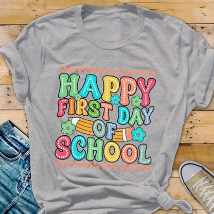 May include: A light gray short-sleeve t-shirt featuring a colorful graphic with the text "HAPPY FIRST DAY OF SCHOOL" in bubble letters. The design includes a yellow pencil illustration, small teal flowers, and orange checkered borders.
