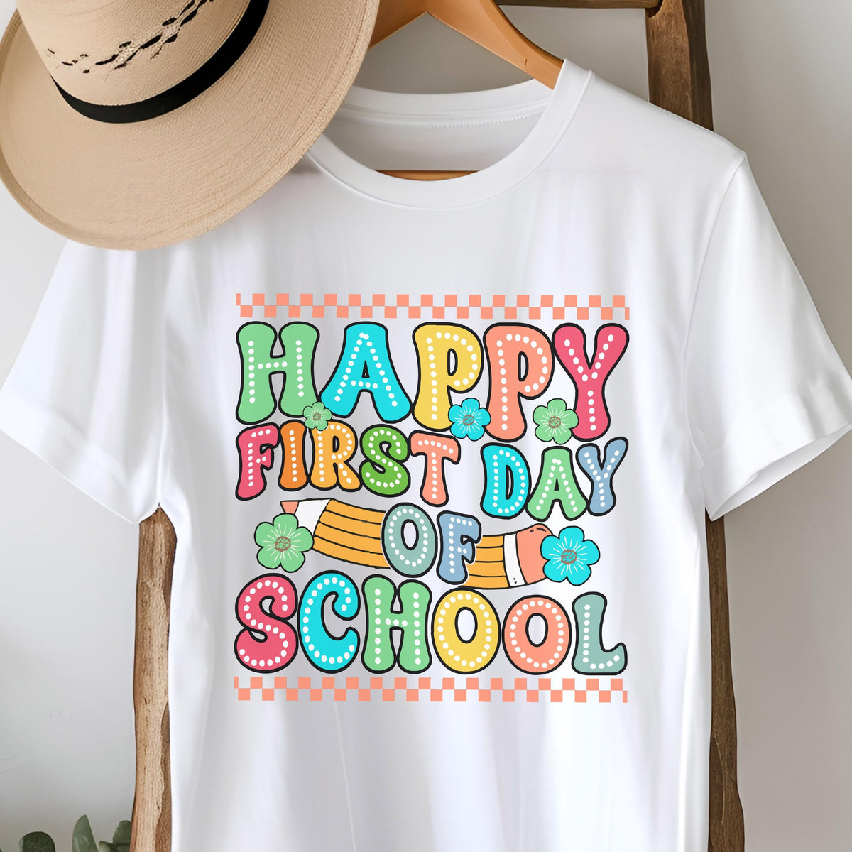 May include: A white t-shirt featuring a colorful graphic with the text "HAPPY FIRST DAY OF SCHOOL" in a bubble font. The design includes a yellow pencil, small flowers, and a checkered border in orange and white. A tan hat rests above the shirt.