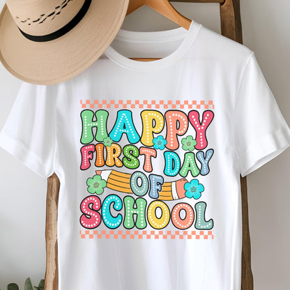 May include: A white t-shirt featuring a colorful graphic with the text "HAPPY FIRST DAY OF SCHOOL" in a bubble font. The design includes a yellow pencil, small flowers, and a checkered border in orange and white. A tan hat rests above the shirt.