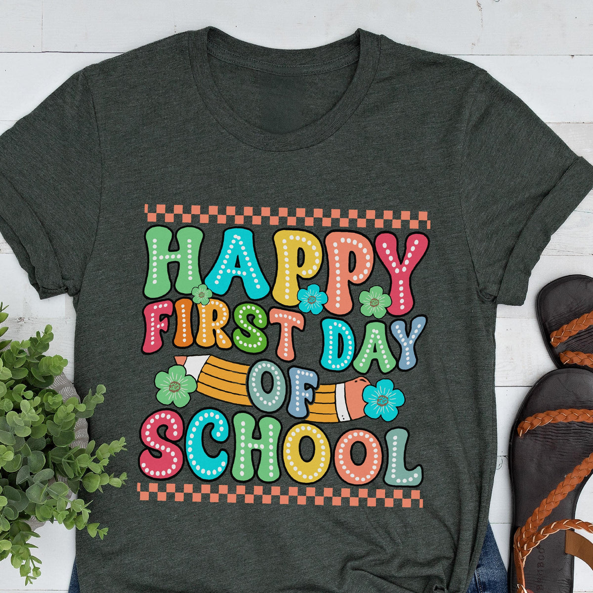 May include: A dark charcoal gray short-sleeve t-shirt featuring a colorful graphic print with the text "HAPPY FIRST DAY OF SCHOOL" in a retro bubble font. The design includes a yellow pencil illustration and small teal and green flowers.