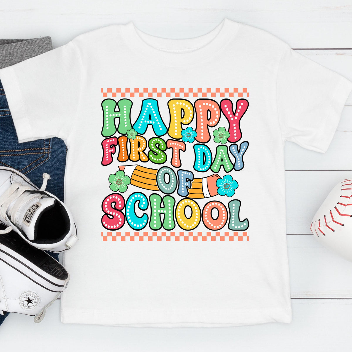 May include: A white short-sleeved t-shirt featuring a colorful graphic print that reads Happy First Day Of School in bubble letters. The design includes a yellow pencil illustration, small blue and green flowers, and orange checkered borders.