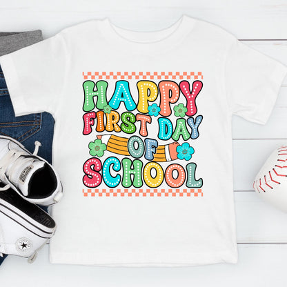 May include: A white short-sleeved t-shirt featuring a colorful graphic print that reads Happy First Day Of School in bubble letters. The design includes a yellow pencil illustration, small blue and green flowers, and orange checkered borders.