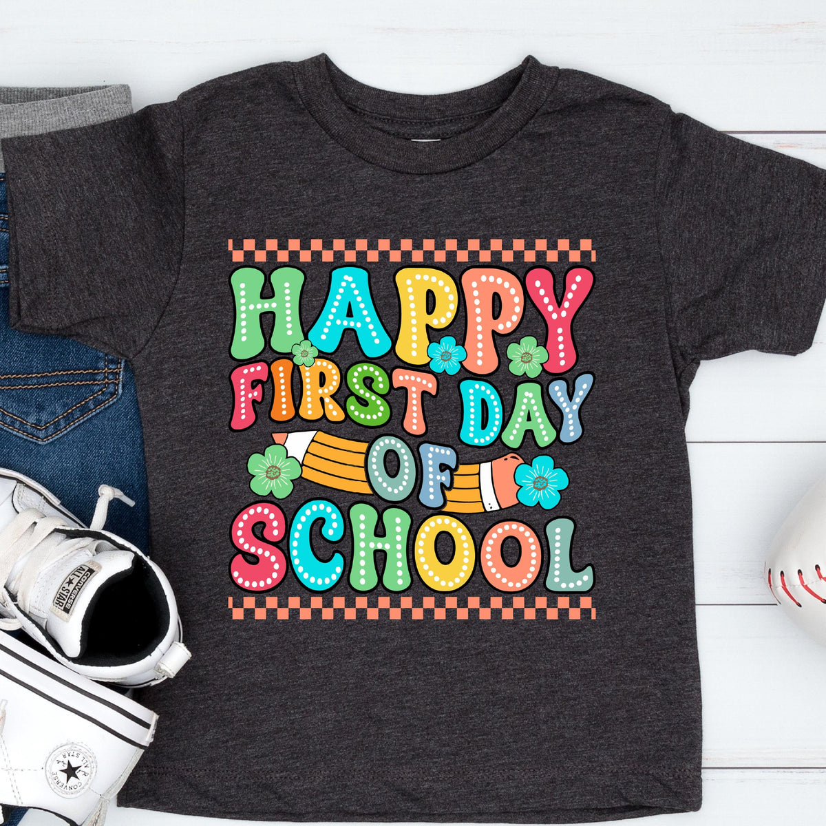 May include: A dark charcoal gray short-sleeve t-shirt featuring a colorful graphic print that reads "HAPPY FIRST DAY OF SCHOOL" in bold, dotted letters. The design includes a yellow pencil illustration and small teal and green flowers.