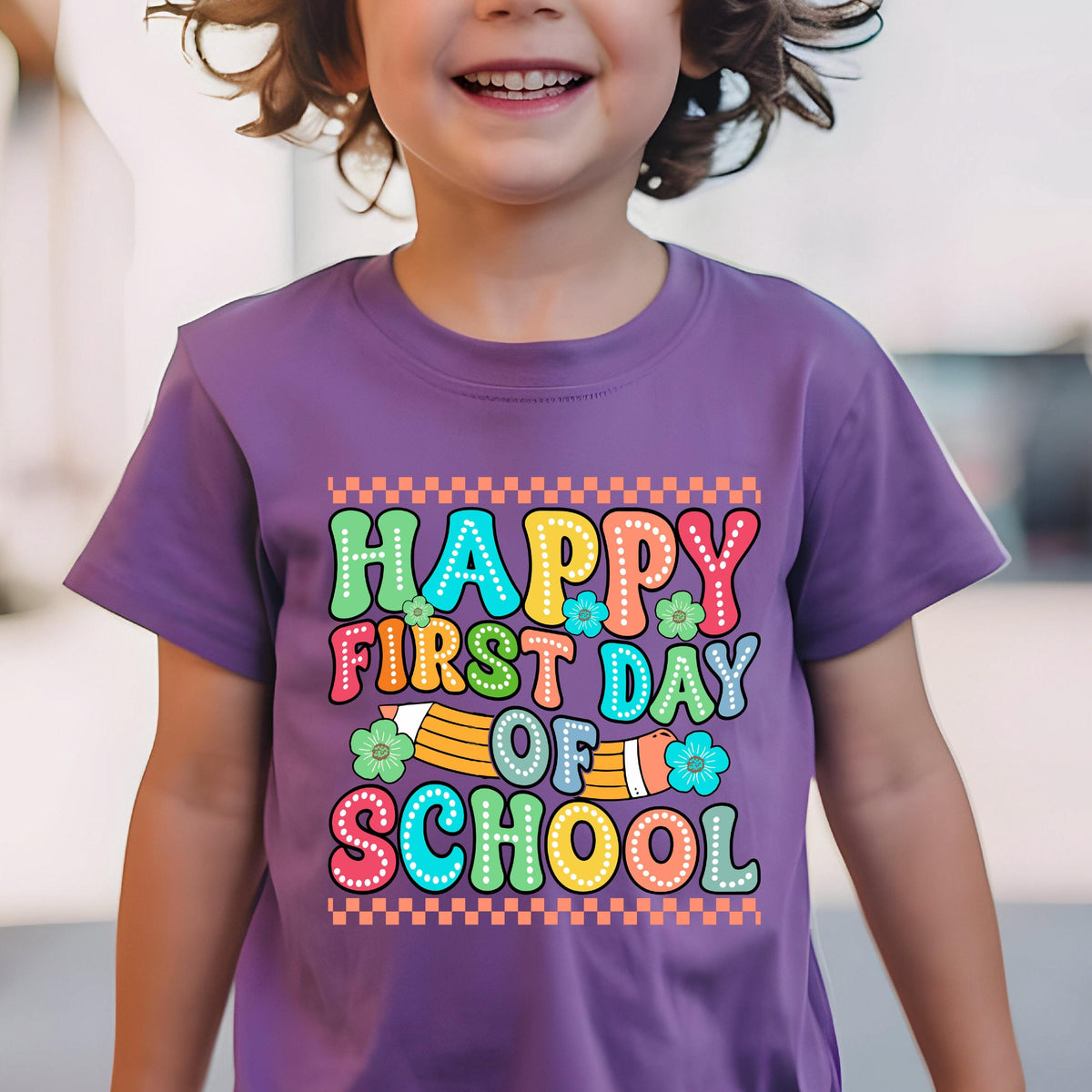 May include: A purple short-sleeved t-shirt featuring a graphic print with the text "HAPPY FIRST DAY OF SCHOOL" in colorful, dotted bubble letters. The design includes a yellow pencil illustration, small flowers, and an orange checkered border.