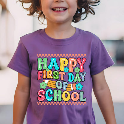May include: A purple short-sleeved t-shirt featuring a graphic print with the text "HAPPY FIRST DAY OF SCHOOL" in colorful, dotted bubble letters. The design includes a yellow pencil illustration, small flowers, and an orange checkered border.