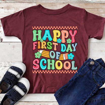 May include: A maroon short-sleeved t-shirt featuring a graphic with the text "HAPPY FIRST DAY OF SCHOOL" in colorful, dotted bubble letters. The design includes a yellow pencil, small teal flowers, and a checkered border on a maroon background.