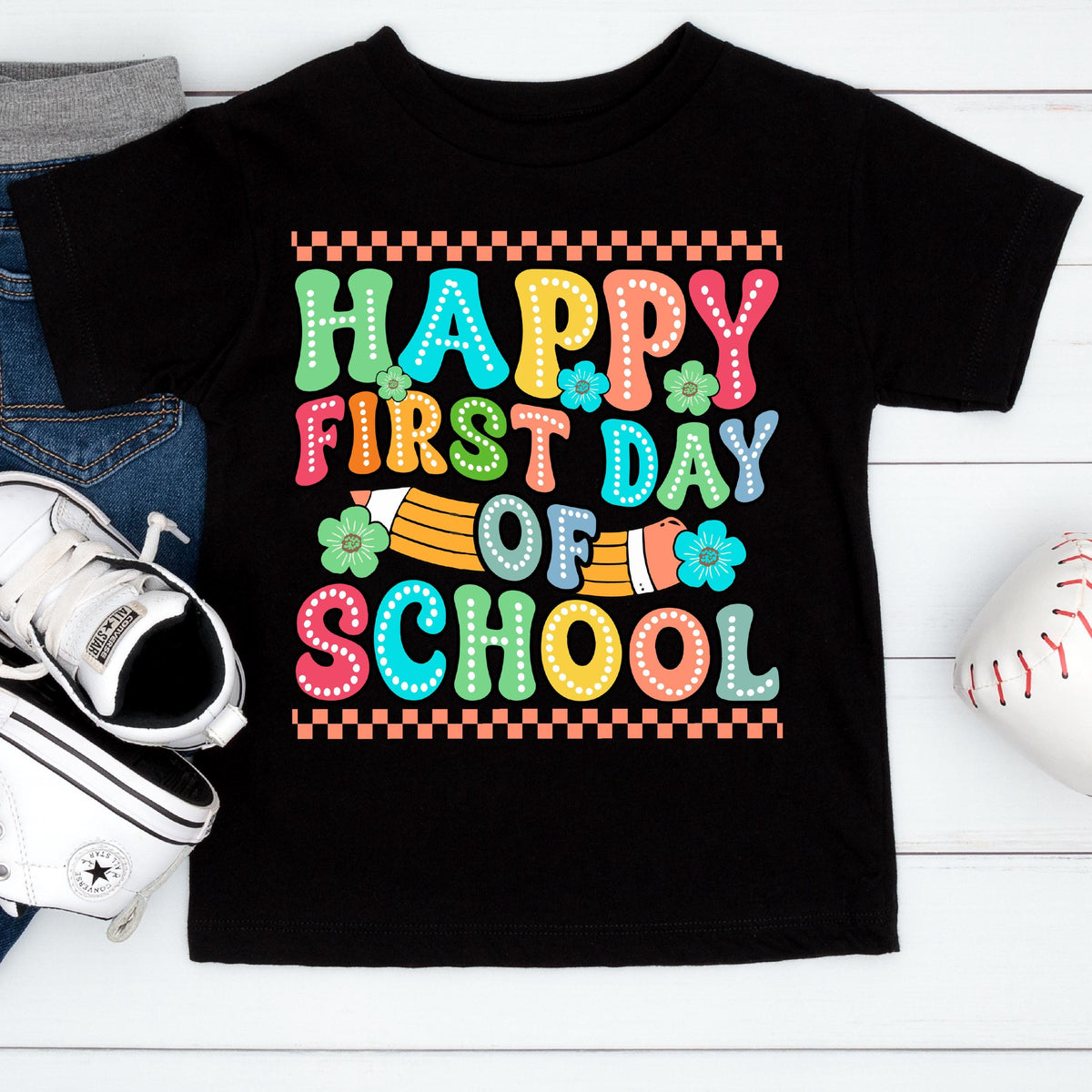 May include: A black short-sleeve t-shirt featuring a colorful graphic with the text "HAPPY FIRST DAY OF SCHOOL" in a bubbly, dotted font. The design includes a yellow pencil illustration, small teal flowers, and a checkered border pattern.