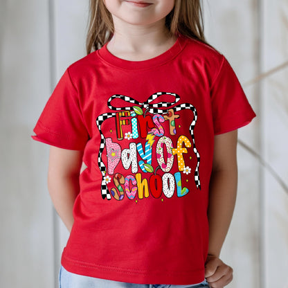 May include: A red t-shirt with the words "First Day of School" in colorful, playful lettering. The text is framed by a black and white checkered ribbon design. The shirt is a short-sleeved style.