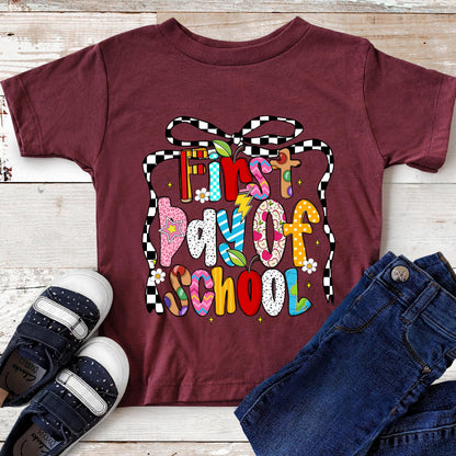 May include: Burgundy short-sleeved t-shirt with the text "First Day of School" in colorful, patterned letters. The design includes a black and white checkered bow. A pair of navy blue shoes and blue jeans are next to the shirt.