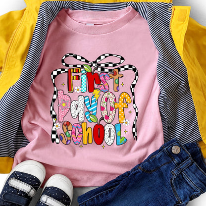 May include: A pink t-shirt with the text "First Day of School" in colorful, patterned letters. The shirt is paired with blue jeans, navy sneakers, and a yellow jacket with black and white striped lining. The design includes a black and white checkered bow.