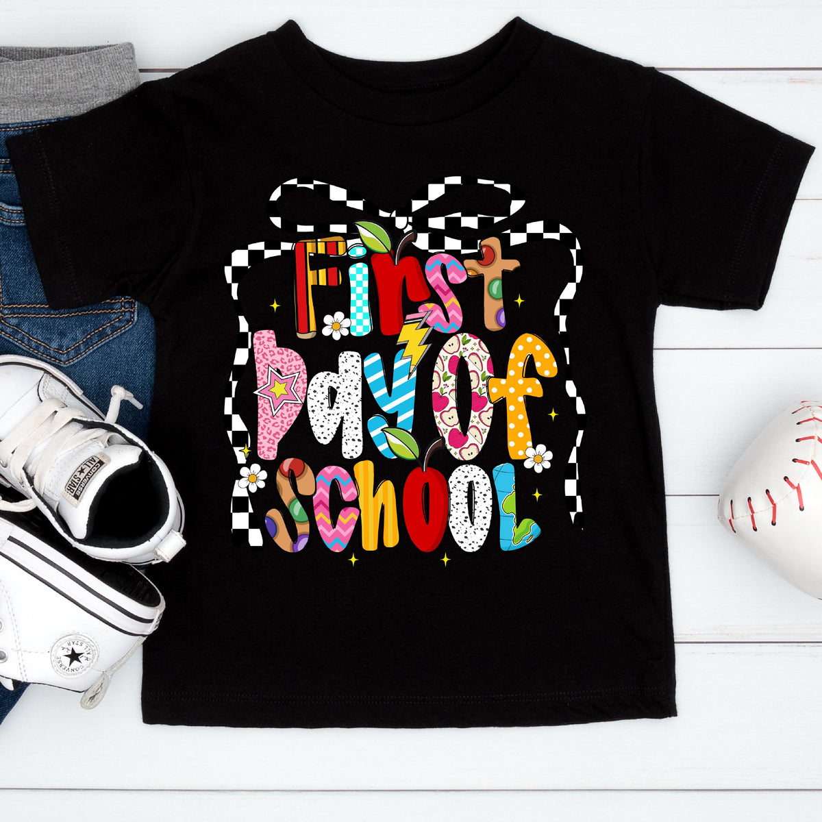 May include: Black t-shirt with the words "First Day of School" in colorful, patterned letters. The design includes stars, flowers, and a checkered ribbon. A pair of white sneakers and a baseball are visible.