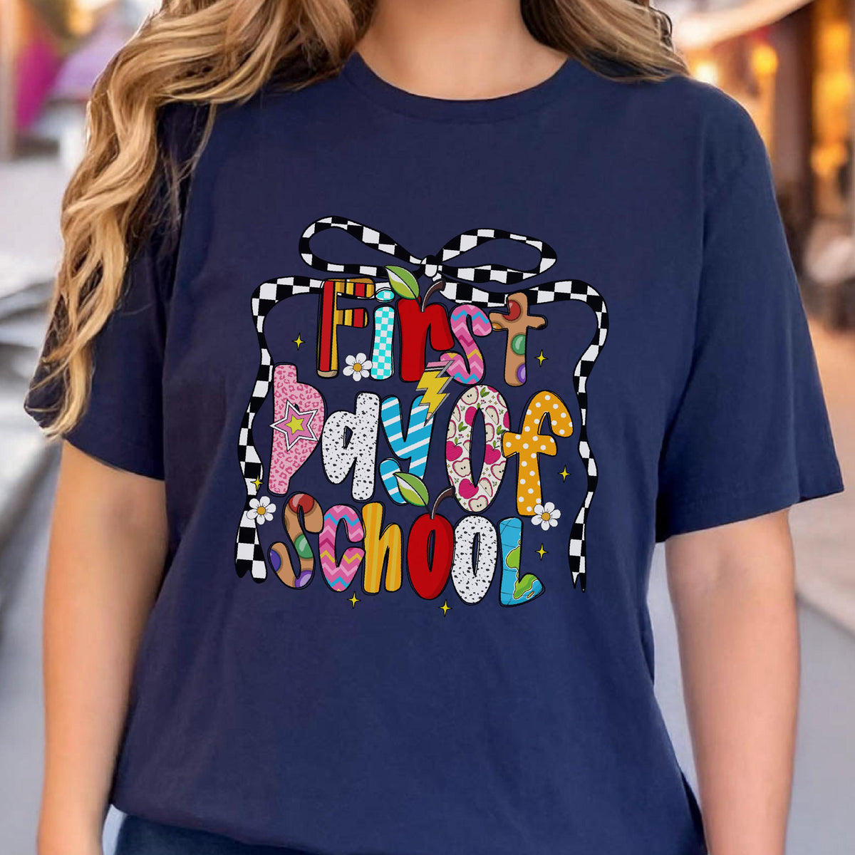 May include: A navy blue short-sleeved t-shirt featuring a graphic print of a black and white checkered bow. The text "First Day of School" is displayed in colorful, patterned letters, accented with small apple, star, and flower illustrations.