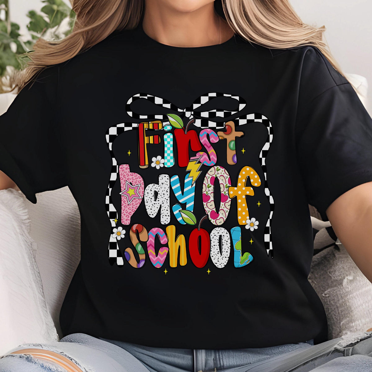 May include: A black short-sleeved t-shirt featuring a colorful graphic with the text First Day of School. The design is framed by a black and white checkered ribbon and includes various patterns, small flowers, a lightning bolt, and apple icons.