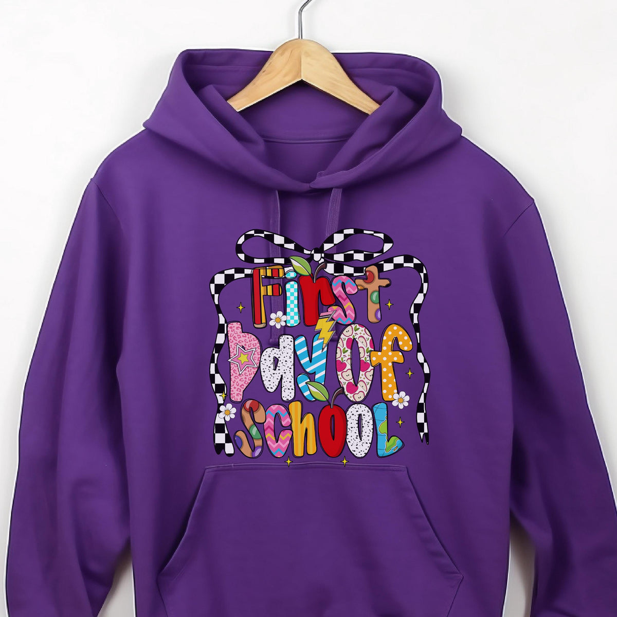 May include: A purple hooded sweatshirt featuring a graphic print with the text First Day of School in colorful, patterned letters. The design is framed by a black and white checkered ribbon and includes small flower and apple illustrations.