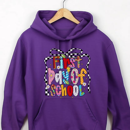 May include: A purple hooded sweatshirt featuring a graphic print with the text First Day of School in colorful, patterned letters. The design is framed by a black and white checkered ribbon and includes small flower and apple illustrations.