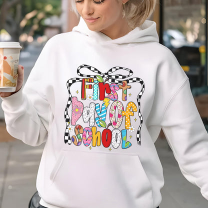 May include: A white hooded sweatshirt featuring a colorful graphic on the front with the text First Day of School. The design includes a black and white checkered bow outline, small flowers, and letters filled with various patterns like polka dots and stripes.
