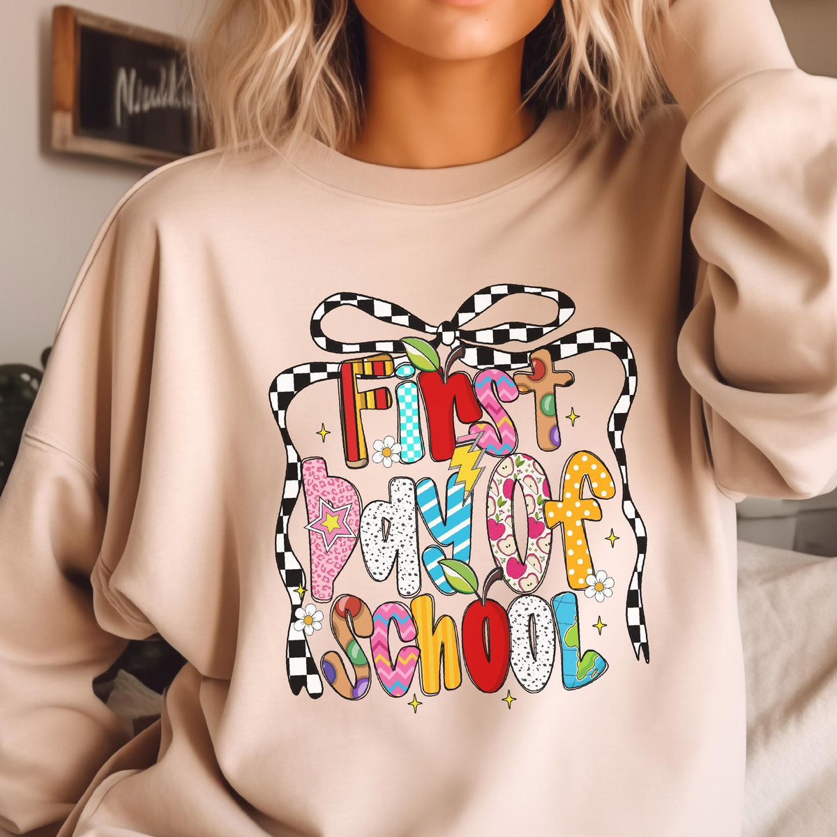 May include: A beige crewneck sweatshirt featuring a colorful graphic print that reads First day of SCHOOL. The text is decorated with various patterns, small apples, and a large black and white checkered bow tied at the top of the design.