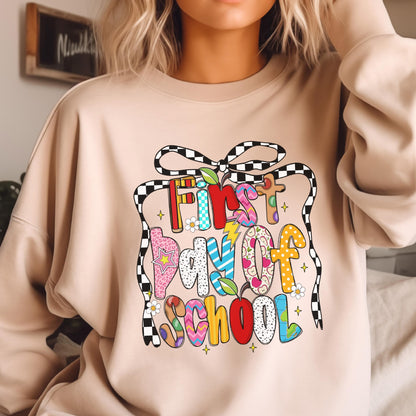 May include: A beige crewneck sweatshirt featuring a colorful graphic print that reads First day of SCHOOL. The text is decorated with various patterns, small apples, and a large black and white checkered bow tied at the top of the design.