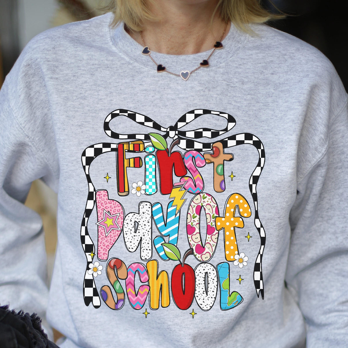 May include: A light gray crewneck sweatshirt featuring a colorful graphic print that reads First day of School in multi-patterned, vibrant letters. The design includes a black and white checkered bow, small white flowers, and red apple icons.