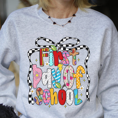 May include: A light gray crewneck sweatshirt featuring a colorful graphic print that reads First day of School in multi-patterned, vibrant letters. The design includes a black and white checkered bow, small white flowers, and red apple icons.