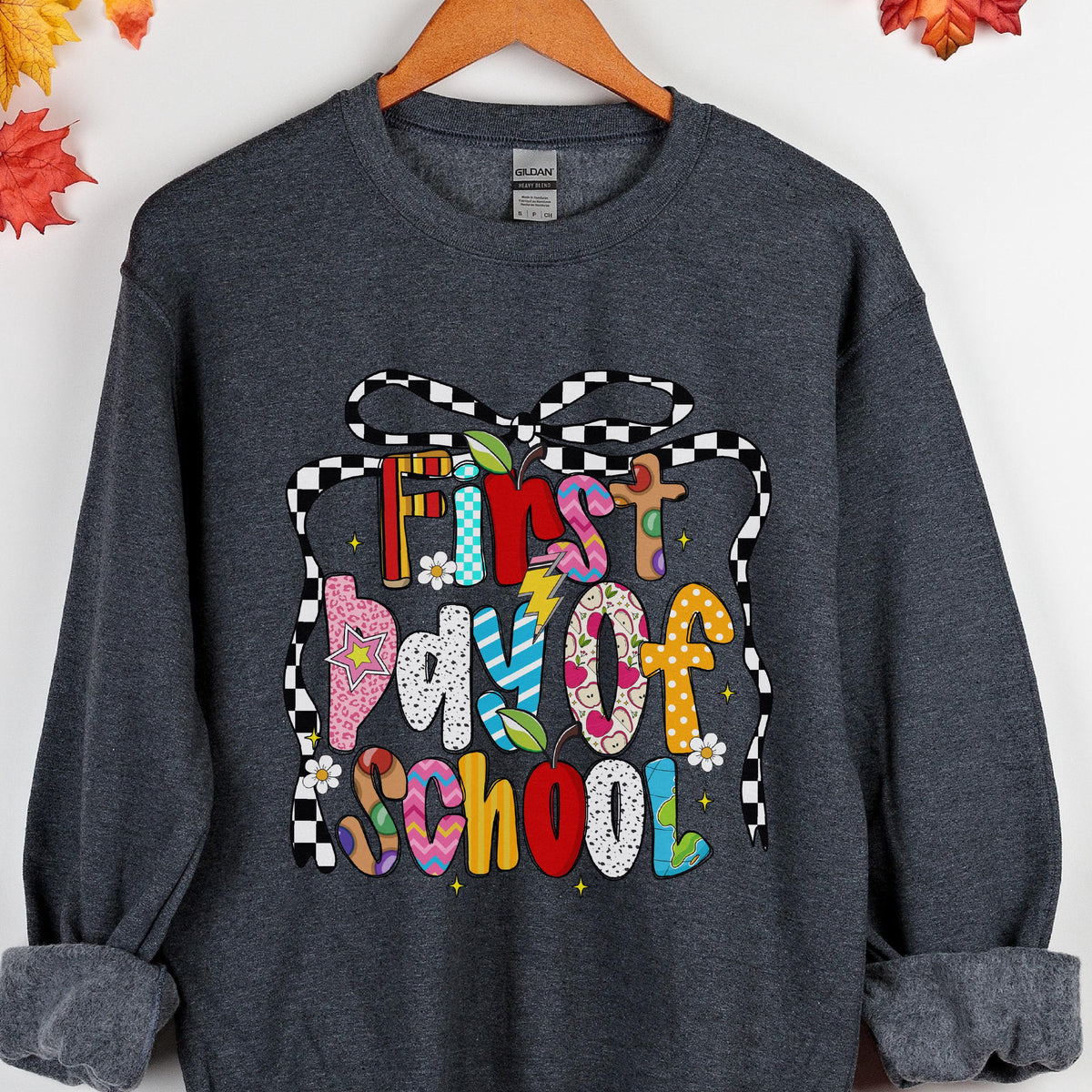 May include: A dark heather gray long-sleeve sweatshirt featuring a graphic print with the text First Day of School. The letters are filled with various colorful patterns including polka dots, stripes, and stars, framed by a black and white checkered bow.