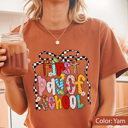 May include: A yam-colored t-shirt with a colorful graphic that reads "First Day of School." The design features a gift box shape with a checkered ribbon and various decorative elements, including flowers and stars. The text is in a playful, multi-colored font.