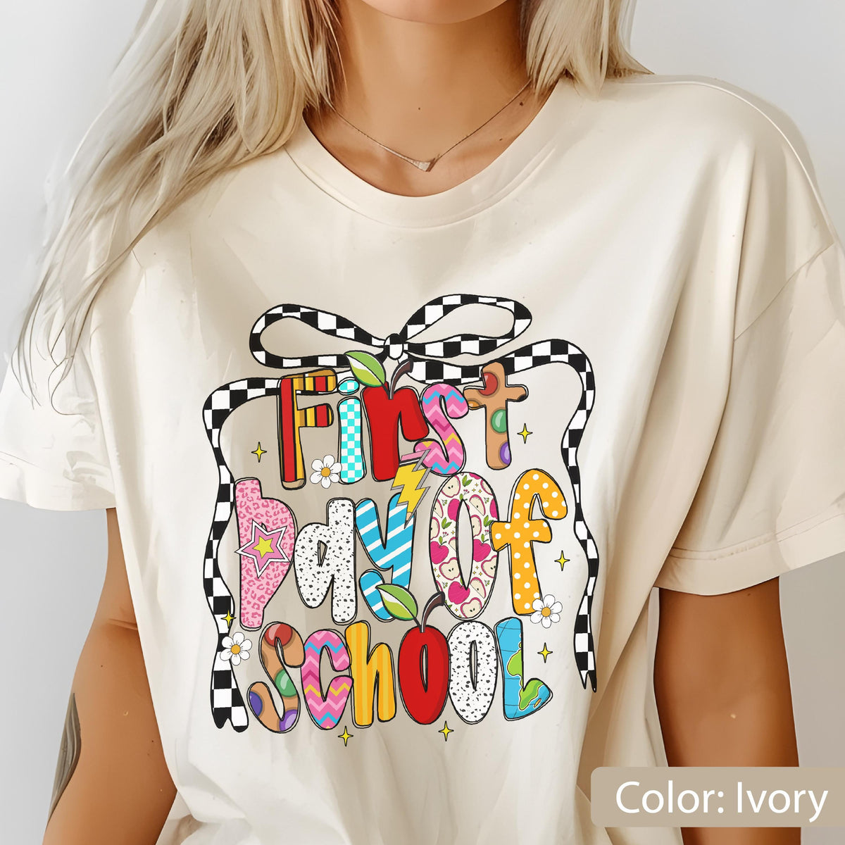 May include: Ivory-colored t-shirt with a colorful "First Day of School" graphic. The text is in various patterns and colors, including red, blue, and yellow. A black and white checkered ribbon design surrounds the text. The shirt is a casual, short-sleeved style.