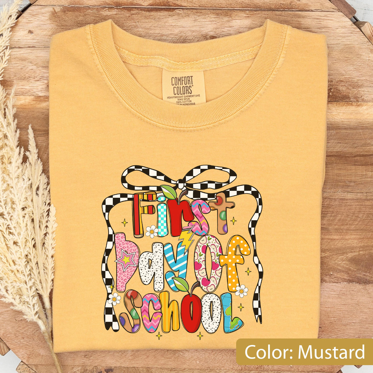 May include: Mustard-colored t-shirt with a colorful "First Day of School" graphic. The graphic features a gift box design with a checkered ribbon and various decorative elements. The text is in a playful, multi-colored font.