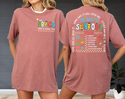 May include: A dusty rose-colored t-shirt with a white collar. The front features the text "THE AB7CD BACK TO SCHOOL TOUR". The back of the shirt has the text "THE BACK TO SCHOOL TOUR" with a list of school-related activities.