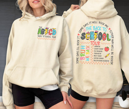 May include: A cream-colored hoodie with a front pocket and a hood. The front features the text "THE ABCD BACK TO SCHOOL TOUR" in colorful letters. The back displays "THE BACK TO SCHZOOL TOUR" with a list of school-related tasks and the phrase "SOME DAYS I'LL ROCK IT".