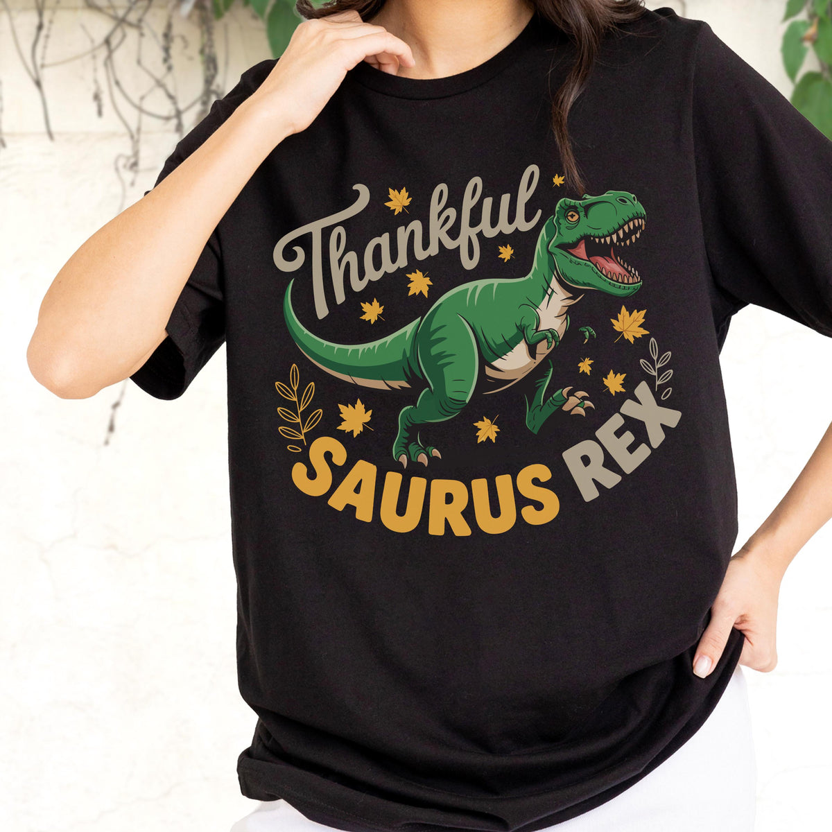 May include: Black t-shirt featuring a green Tyrannosaurus Rex illustration with the text "Thankful Saurus Rex" in a playful font. The design includes autumn leaves and stars, perfect for a Thanksgiving or dinosaur-themed gift.