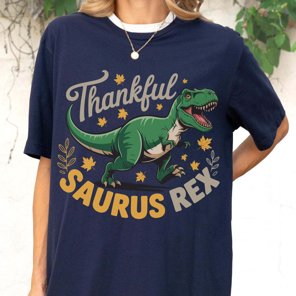 May include: Navy blue t-shirt with a white collar featuring a green Tyrannosaurus Rex illustration. The design includes the text "Thankful Saurus Rex" in gray and yellow, with autumn leaf accents. A casual, graphic tee.