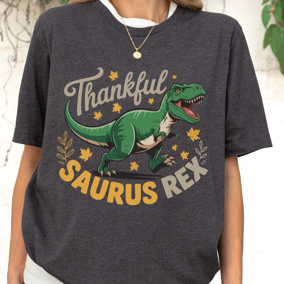 May include: Dark gray t-shirt with a graphic of a green Tyrannosaurus Rex and the words "Thankful Saurus Rex" in a playful font. The design includes yellow maple leaves and decorative elements.