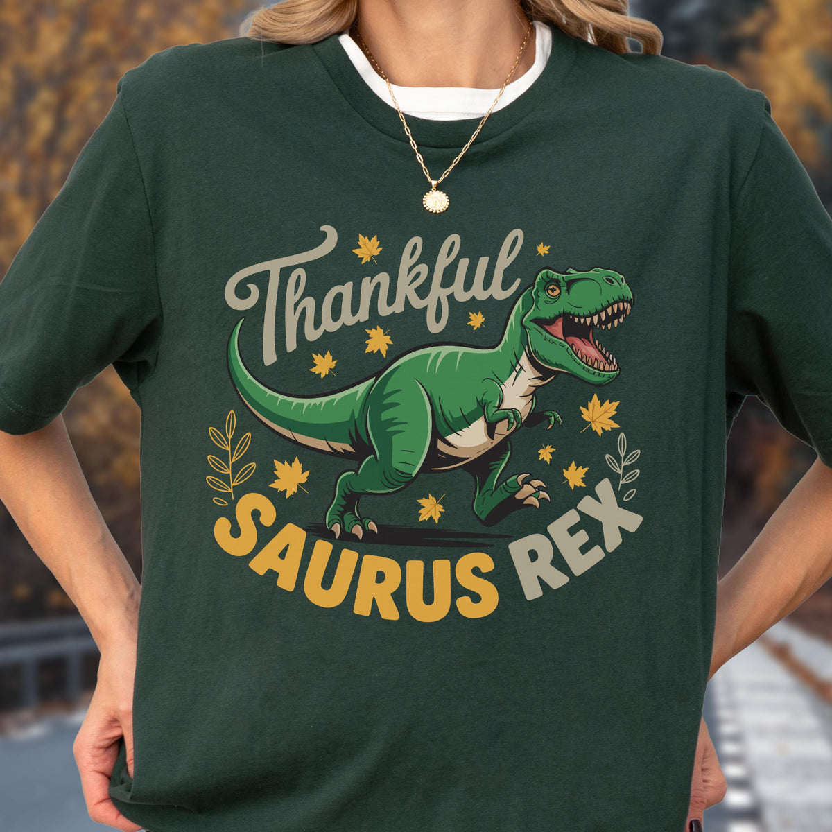 May include: Dark green t-shirt featuring a green Tyrannosaurus Rex illustration with the text "Thankful Saurus Rex" in a playful font. The design includes yellow maple leaves and decorative elements.