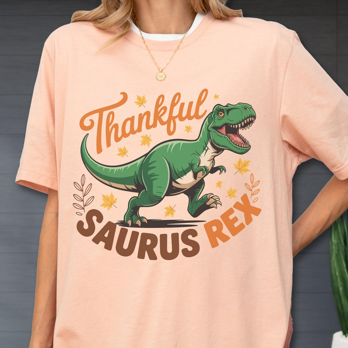 May include: A peach-colored t-shirt featuring a green Tyrannosaurus Rex dinosaur with the text "Thankful Saurus Rex" in orange and brown. The design includes fall leaves and decorative elements, creating a festive and playful look.
