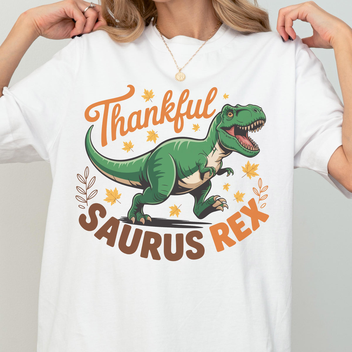 May include: White t-shirt featuring a green Tyrannosaurus Rex illustration with the text "Thankful Saurus Rex" in orange and brown. The design includes fall leaves and decorative elements, perfect for Thanksgiving.
