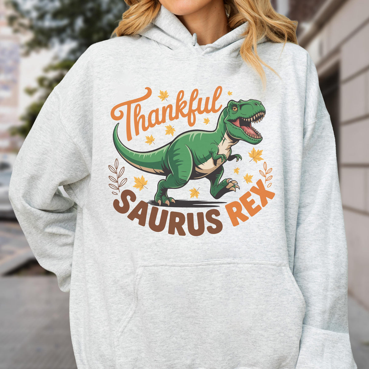 May include: A light gray hoodie featuring a green Tyrannosaurus Rex illustration with the words "Thankful Saurus Rex" in orange and brown. The design includes fall leaves and a decorative border.