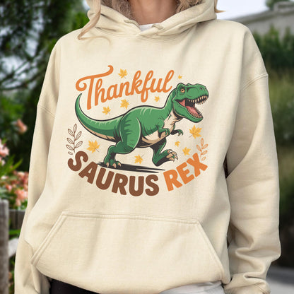 May include: A beige hoodie featuring a green Tyrannosaurus Rex illustration with the text "Thankful Saurus Rex" in orange and brown. The design includes fall leaves and decorative elements, perfect for autumn.