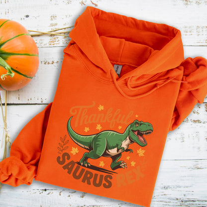 May include: An orange hoodie featuring a green cartoon Tyrannosaurus Rex with the words "Thankful Saurus Rex". The design includes fall leaves. A pumpkin is visible in the background. The hoodie is made of a soft material.