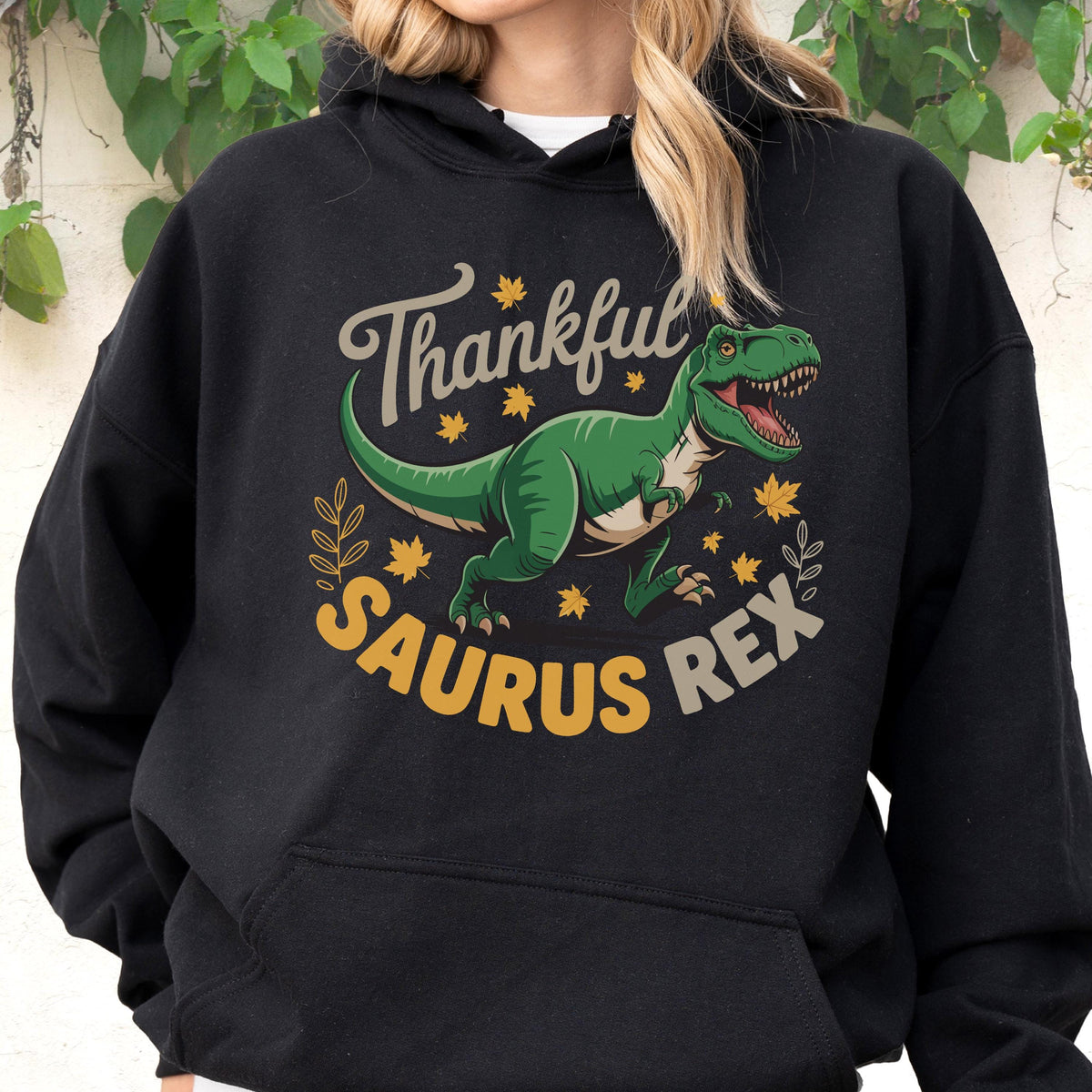 May include: Black hooded sweatshirt with a graphic of a green Tyrannosaurus Rex and the words "Thankful Saurus Rex" in a playful font. The design includes fall leaves and decorative elements.