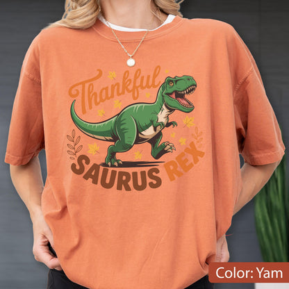 May include: A yam-colored t-shirt with a green Tyrannosaurus Rex graphic. The text reads "Thankful Saurus Rex" in a fall-themed design. The shirt is a short-sleeved crew neck.